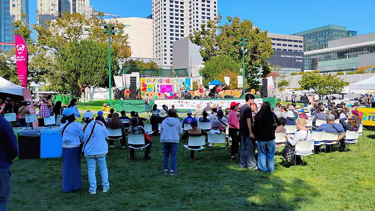 A wide shot of people watching a performance on an outdoor stage in a city park. A wide shot of people watching a performance on an outdoor stage in a city park.