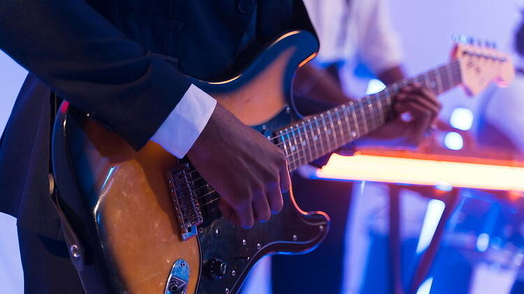 Close-up of hands playing an electric guitar on a stage with colorful lights in the background.