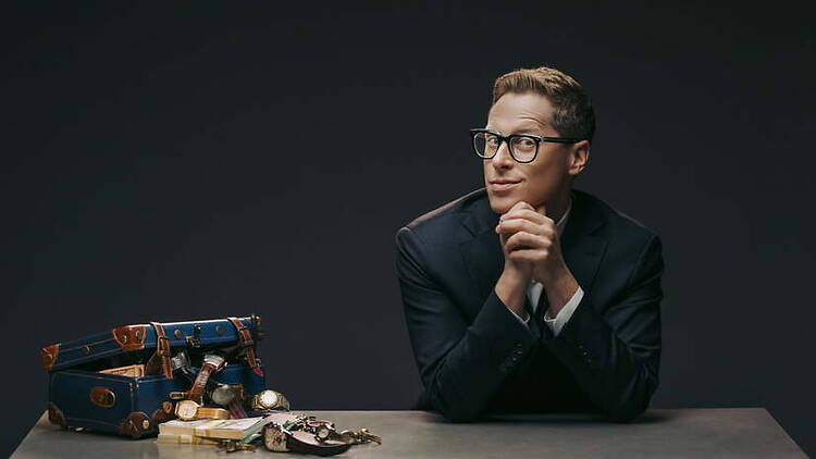 A wide shot of a man in a suit posing beside vintage luggage and watches on a table.
