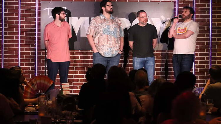 Wide shot of four people speaking on a comedy club stage before an audience. Wide shot of four people speaking on a comedy club stage before an audience.