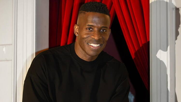 A close-up of a person smiling in a doorway with red curtains.