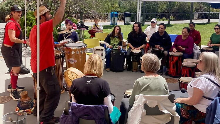 A wide shot of people playing drums together under a canopy outdoors.