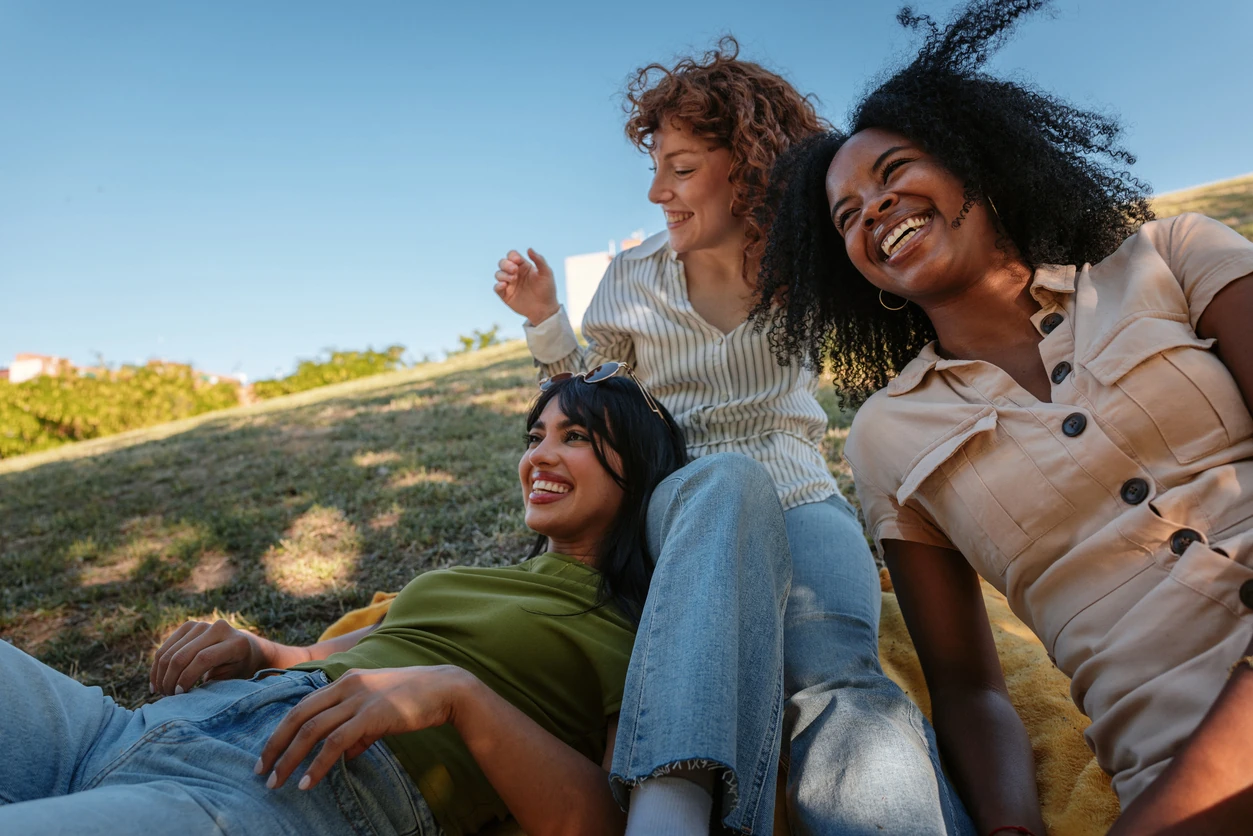 Three friends laughing and enjoying a delightful picnic under the warm sun in a vibrant park, surrounded by nature and greenery