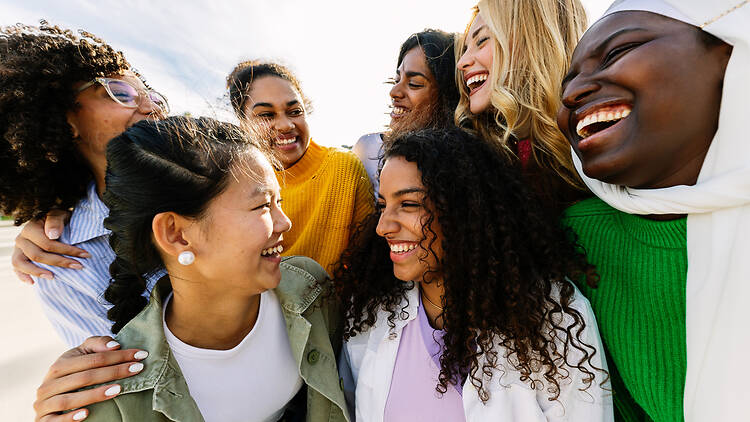 Seven happy young adult women from different cultures laughing together outdoors Seven happy young adult women from different cultures laughing together outdoors. Female friendship concept with diverse group of girls friends hugging each other having fun at city street