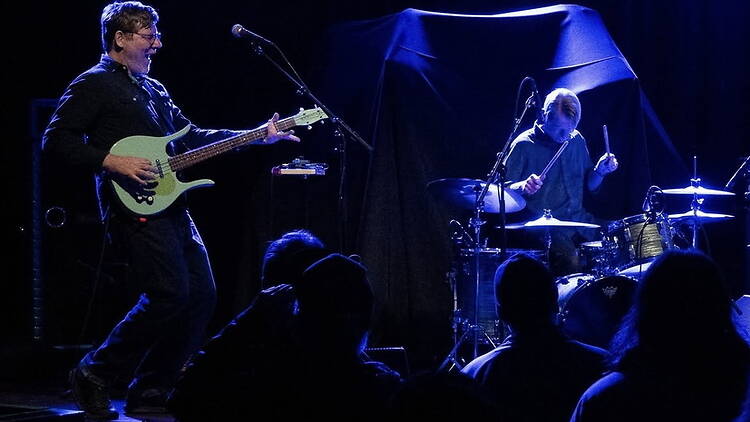 A wide shot of a bassist and drummer performing on stage before a live audience.