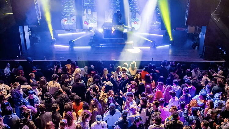 Wide shot of a crowd gathered before a DJ performing on a brightly lit indoor concert stage.
