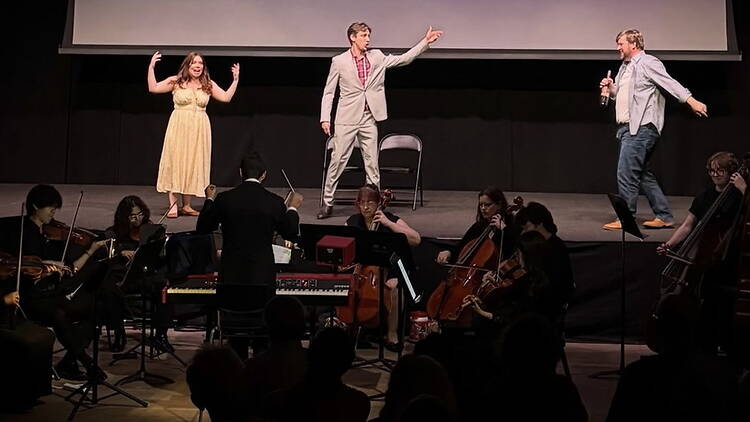 A wide shot of performers and an orchestra performing on a theater stage.