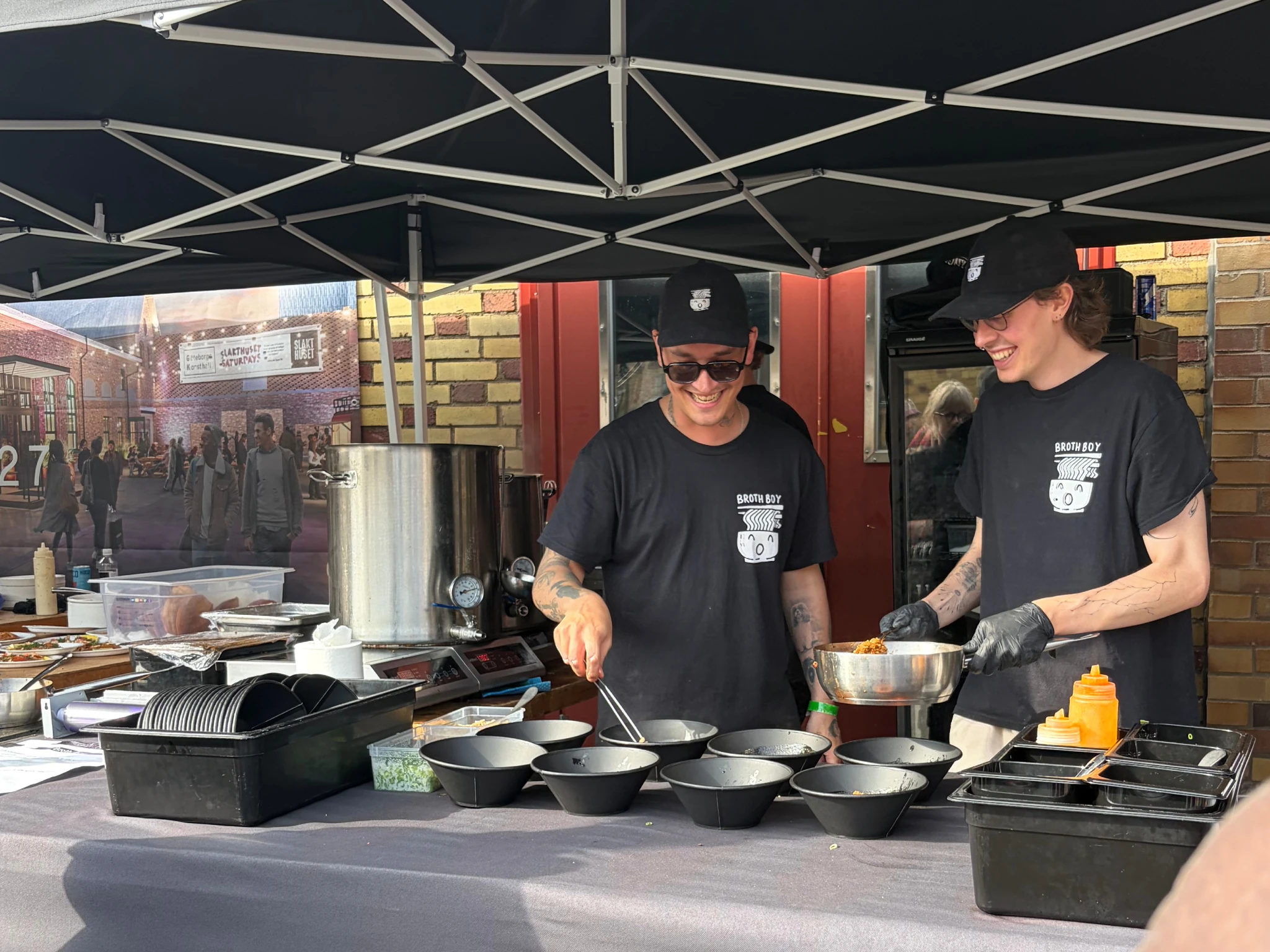 Two smiling vendors in matching Broth Boy black t-shirts preparing bowls of ramen at an outdoor food festival stand in Gothenburg.