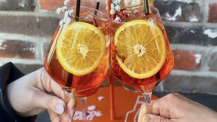 Close-up of two orange drinks being toasted against a brick wall.