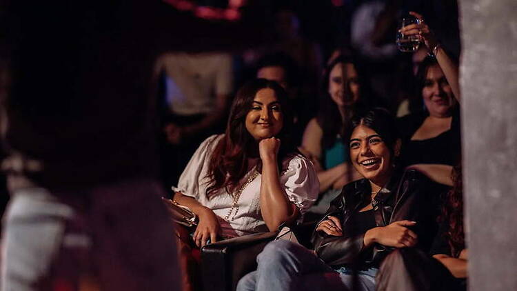 A wide shot of attendees smiling and watching a performance in a dimly lit venue A wide shot of attendees smiling and watching a performance in a dimly lit venue