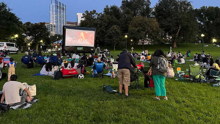 A wide shot of people watching a movie on a large screen in a park
