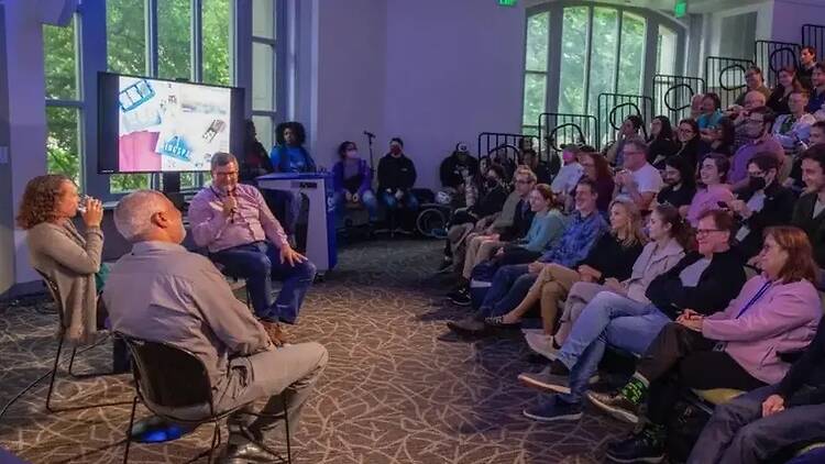 A wide shot of speakers presenting to an audience in an indoor event space. A wide shot of speakers presenting to an audience in an indoor event space.