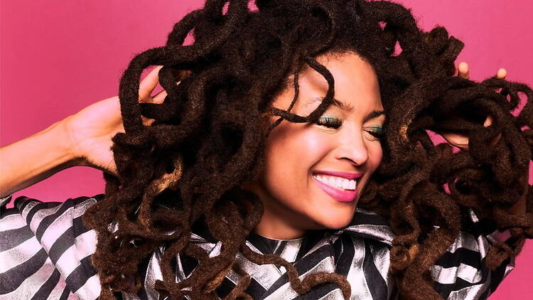 Close-up of a smiling woman with long dreadlocks posing against a pink background.