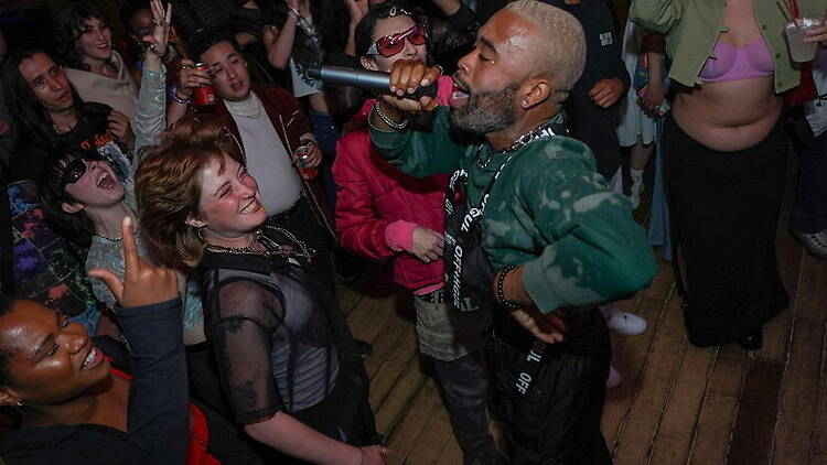A wide shot of a performer singing into a microphone as attendees dance around him indoors