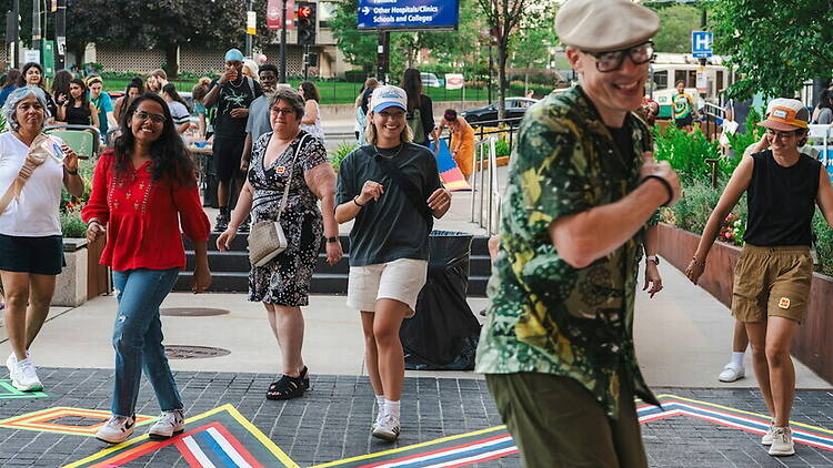 A wide shot of people walking together along a colorful street during an outdoor festival A wide shot of people walking together along a colorful street during an outdoor festival