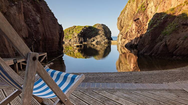 A blue and white striped deck chair overlooking the natural Mermaid Pool cove at Burgh Island, surrounded by steep rocky cliffs.