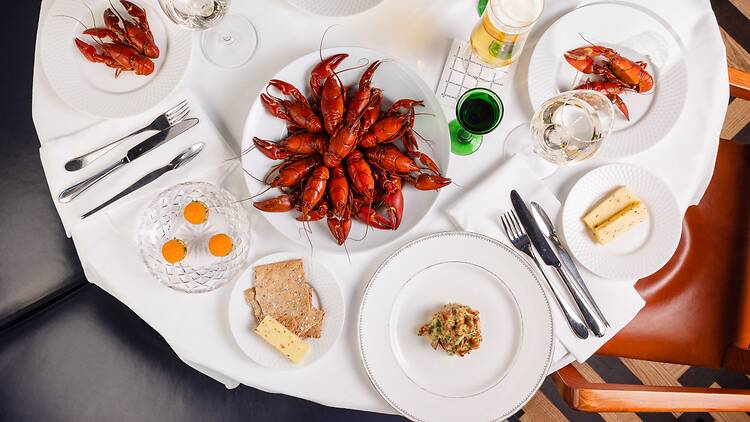 Overhead view of a festive Swedish crayfish party table setting with plates of cooked red crayfish, cheese, crispbread, and drinks.