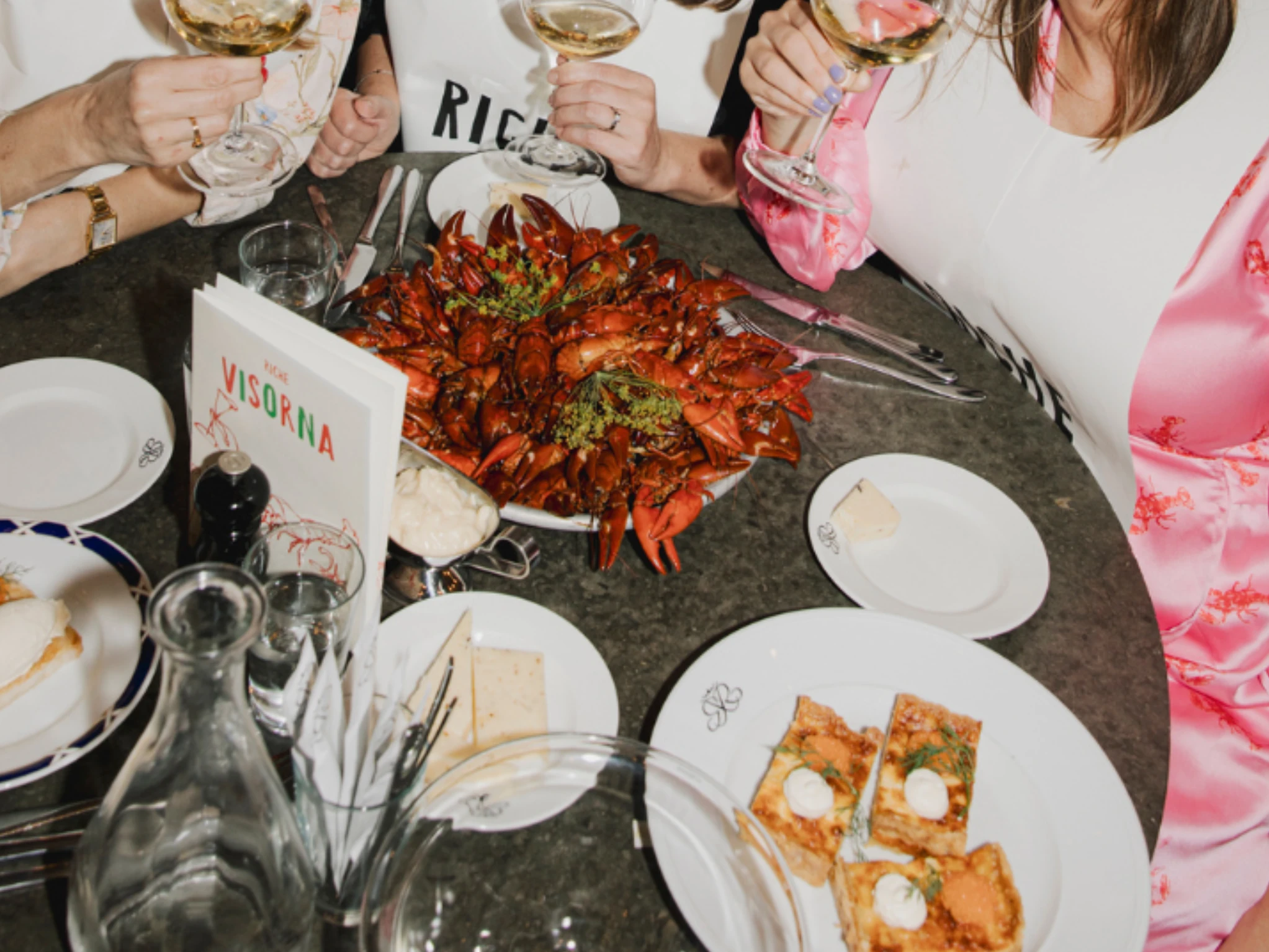 People wearing paper bibs clinking wine glasses over a table filled with red crayfish, V&auml;sterbotten cheese pie, and a songbook at Restaurant Riche.