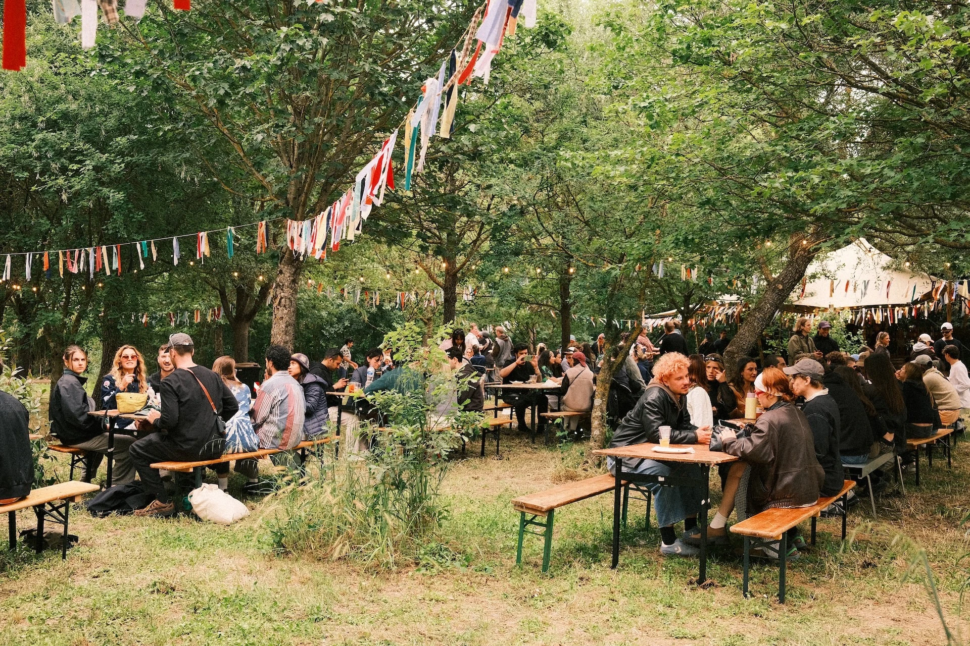 Festival goers sit on long tables at Flash Festival drinking and talking with bunting and tents in the foreground and background