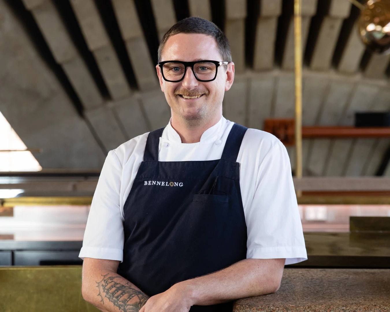 Chef Rob Cockerill, wearing glasses and a black Bennelong apron, smiling inside the restaurant.