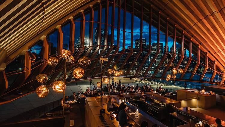 The dining room at Bennelong The interior of the Bennelong dining room, with concrete beams, glass-bulb lighting and views of the Sydney Harbour Bridge.