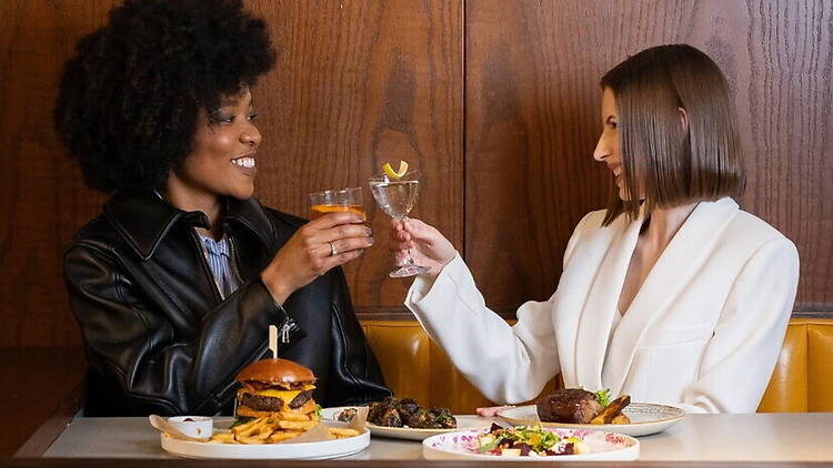 A close-up of two women toast with drinks while dining together at a restaurant.
