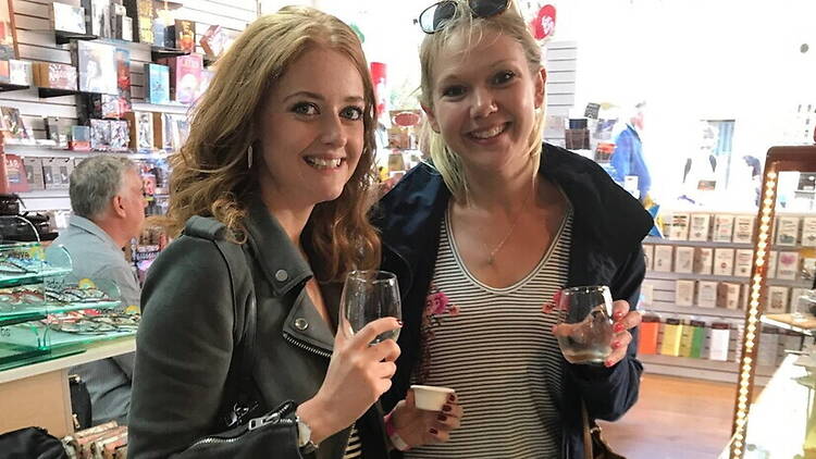A wide shot of two women hold drinks and smile together inside a shop.