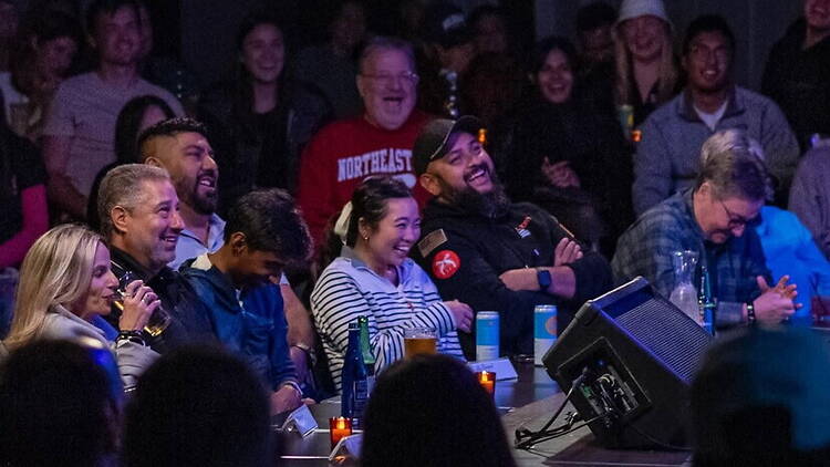 A wide shot of an audience laughing and enjoying a live performance at a venue. A wide shot of an audience laughing and enjoying a live performance at a venue.