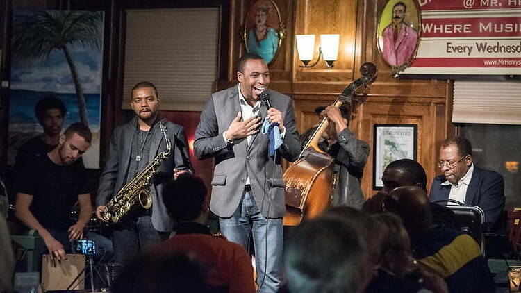 Audience view of a jazz band performing with a vocalist, saxophonist, and bassist onstage at a club.