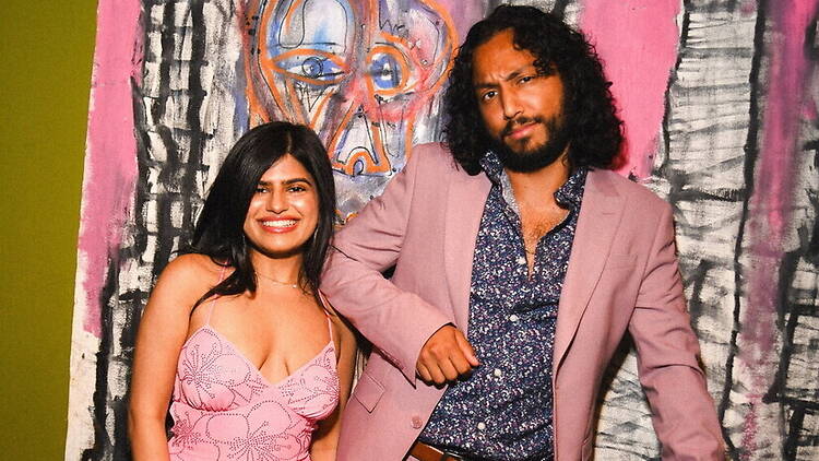A medium shot of two people posing together in front of colorful artwork. A medium shot of two people posing together in front of colorful artwork.