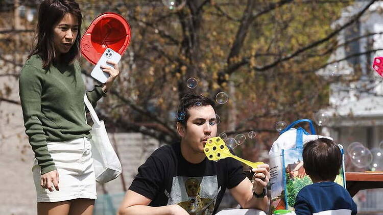 A wide shot of people blowing bubbles with a child outdoors in a park.