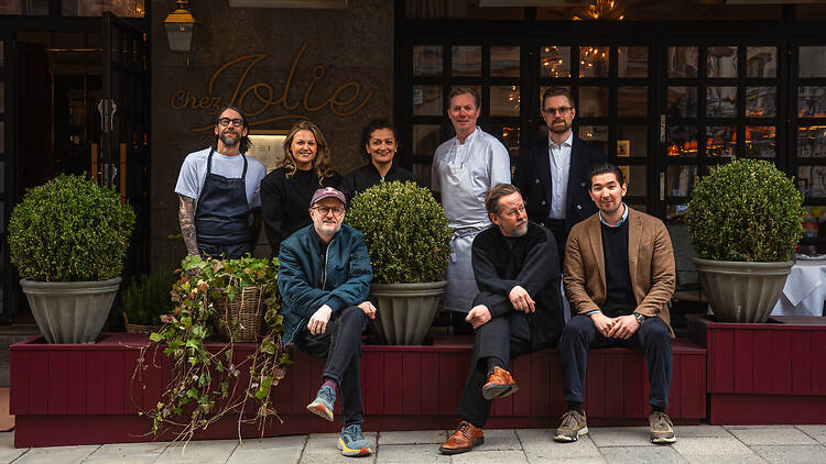 Krogen Chez Jolie på Östermalm Group photo outside Chez Jolie restaurant in Stockholm: eight people, some standing and some seated on a low burgundy planter box, posing together in front of the restaurant's entrance and gold "Chez Jolie" signage, flanked by large potted boxwood shrubs.
