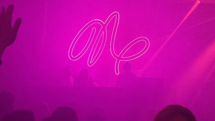 Klubben Natten i Stockholm Neon sign in cursive script glowing white against a magenta-lit club interior, with the silhouettes of a raised hand and the crowd's heads visible in the foreground, and a red laser beam cutting across the upper right corner.