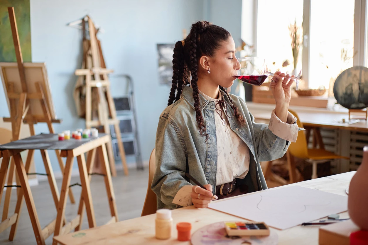 Young adult woman sitting at table drinking red wine while holding paintbrush in art studio, engaging in painting activity with art supplies and canvas visible