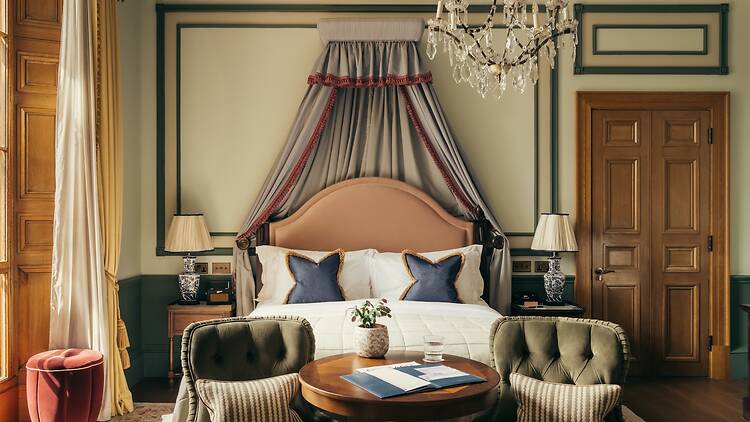 A luxurious, Georgian-style hotel bedroom featuring high ceilings, a draped canopy bed with a pink headboard, a crystal chandelier, two sage-green velvet armchairs at a small round wooden table, and tall wooden sash windows with gold curtains.