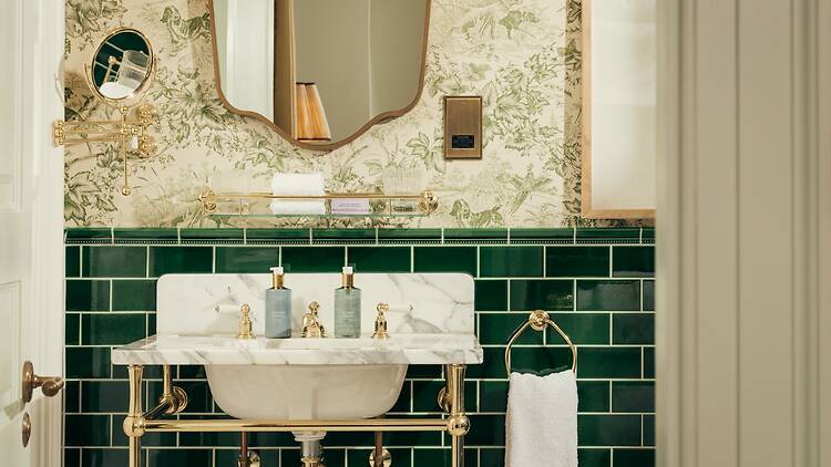 An elegant hotel bathroom featuring a white marble console sink with brass legs, dark green subway-tiled lower walls, green-and-white botanical Toile de Jouy wallpaper, a shaped mirror, and brass fixtures.