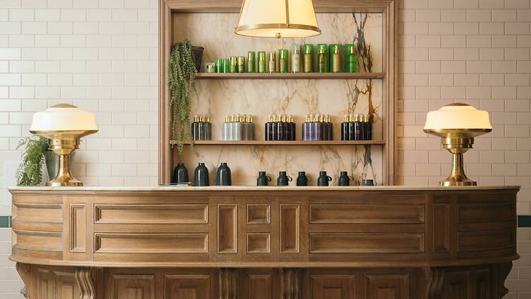 A spa reception desk at Gleneagles Townhouse featuring a curved, paneled oak desk with warm brass banker-style lamps, set against white tiled walls and a marble-lined niche displaying beauty products.