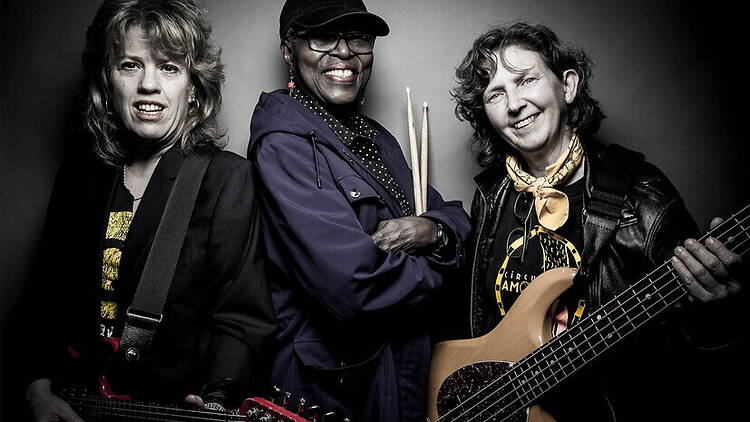 A wide shot of three musicians posing with guitars and drumsticks against a dark backdrop. A wide shot of three musicians posing with guitars and drumsticks against a dark backdrop.