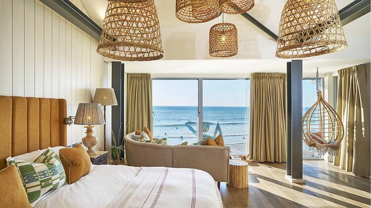 Bright hotel room with woven rattan light fixtures, an orange tufted headboard, a curved sofa, a hanging chair, and floor-to-ceiling windows overlooking an ocean beach.