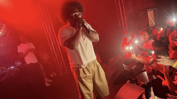 A close-up of a singer performing into a microphone on a red-lit stage before a crowd.