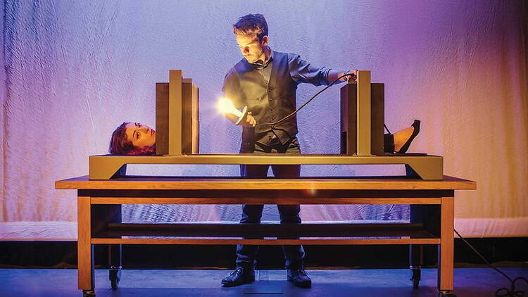 A wide shot of a performer shining a light on a woman inside a table on a stage.