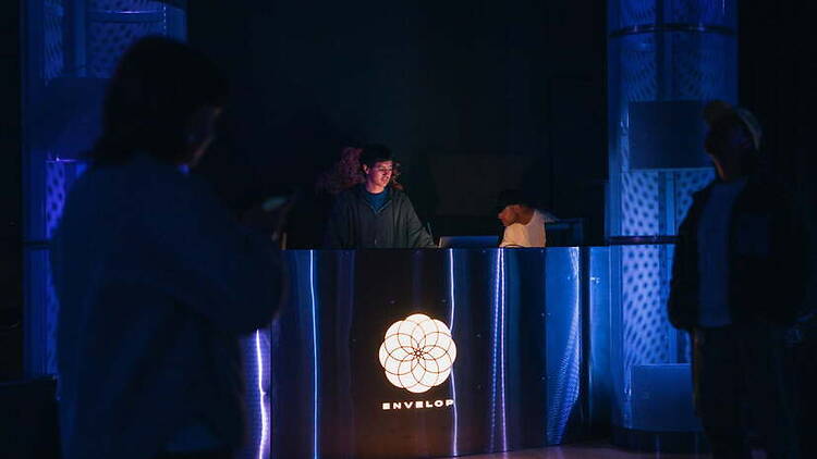A wide shot of a DJ performing at a booth in a dark, blue-lit venue.
