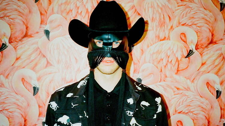 A close-up of a person wearing a cowboy hat and mask against a flamingo-patterned backdrop.