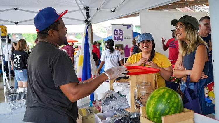 A wide shot of people serving and talking at a food stall under a canopy.