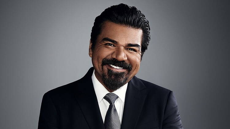 A close-up of a person smiling in a suit and tie against a gray background. A close-up of a person smiling in a suit and tie against a gray background.