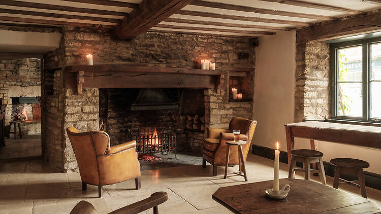 Two leather armchairs facing a glowing open fireplace in a cozy, rustic stone pub room with exposed wooden beams and candlelight.