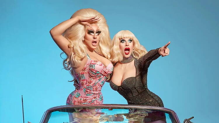 A wide shot of two people posing and pointing from a convertible car against a blue backdrop.