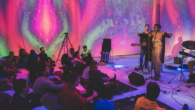 A wide shot of performers singing to an audience in a colorful indoor venue
