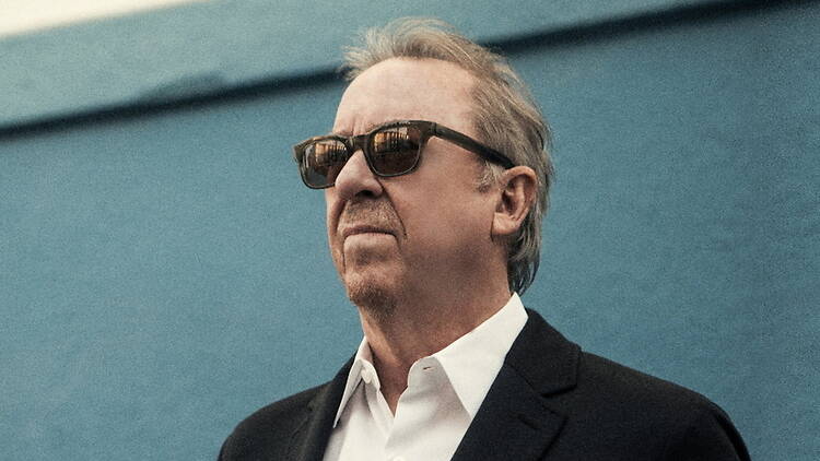 A close-up of a person wearing sunglasses and a black suit against a blue backdrop.