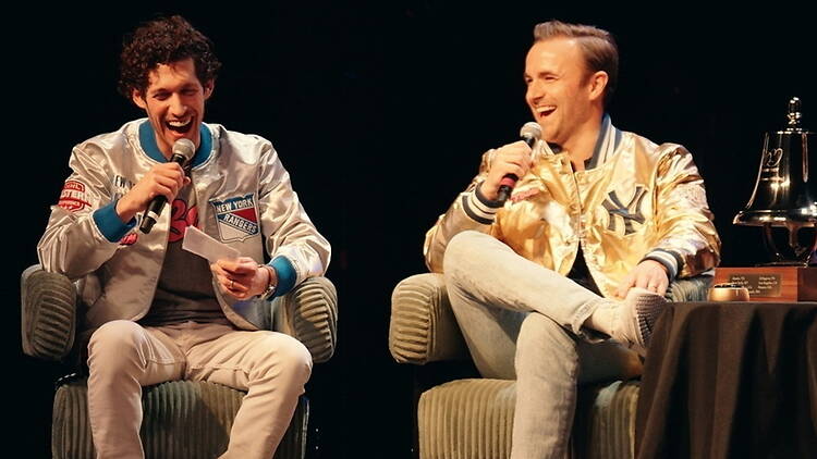 A wide shot of two people speaking into microphones while seated on stage.